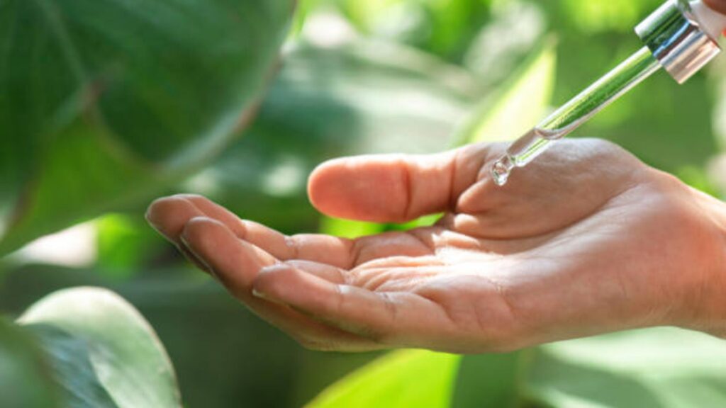 Fondo de plantas verdes con una mano que sostiene una gota de aceite esencial que cae de un gotero.