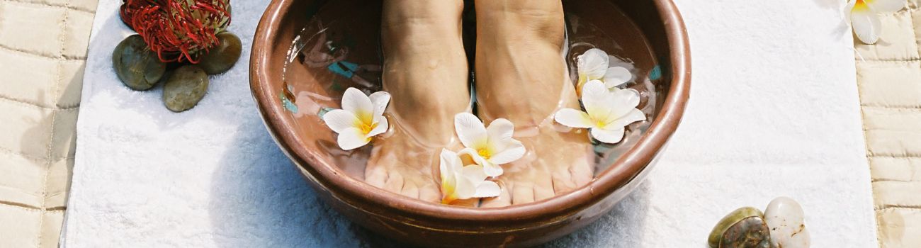 Pies de una mujer caucásica sumergidos en un pequeño cuenco de bambú con agua y flores de jazmín.