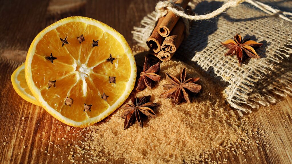 Un trozo de naranja con clavos de olor y ramas de canela puestos en una superficie de madera con anís estrellado a modo decorativo.