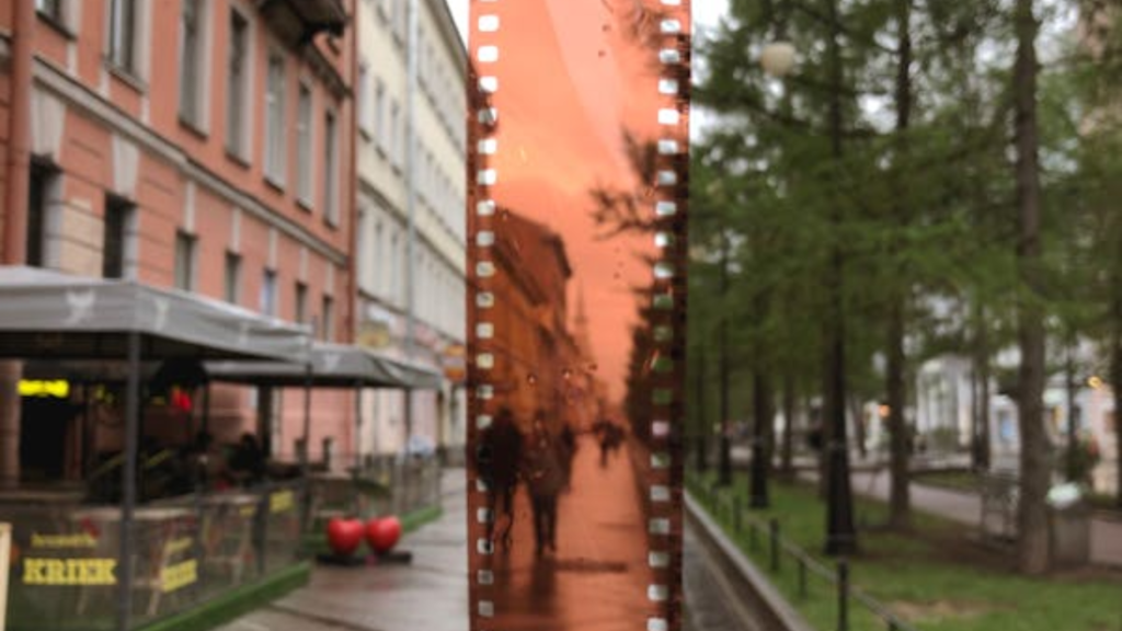 La imagen muestra una escena de una calle capturada a través de una película de cámara fotográfica con gotas de agua en ella. El lado derecho de la imagen está claro y muestra una acera bordeada de árboles, mientras que el lado izquierdo se ve a través de la película mojada, creando un efecto borroso con iluminación naranja.