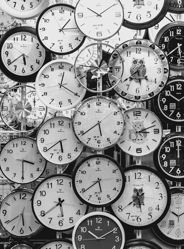 La imagen muestra una colección de varios relojes de pared con diferentes diseños y horas mostradas. Los relojes están en blanco y negro, algunos con números romanos y otros con números estándar. La disposición de los relojes es densa, con algunos superponiéndose a otros, creando un patrón visualmente interesante que llama la atención sobre el concepto del tiempo y su medición.