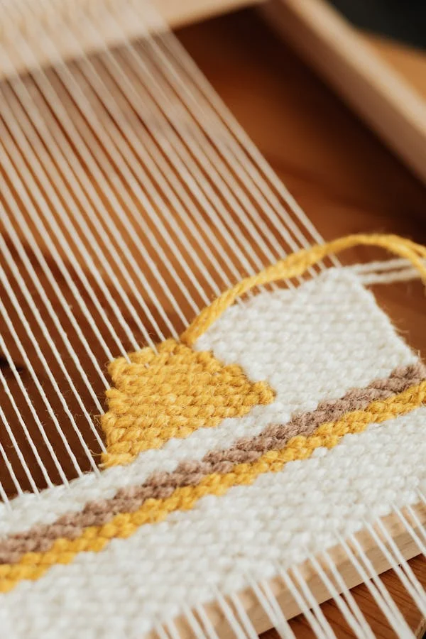 La imagen muestra una vista de cerca de un proyecto de tejido en un telar. El patrón de tejido incluye rayas horizontales en colores blanco, amarillo y marrón. El telar tiene hilos verticales que forman parte de su estructura y mantienen el tejido en su lugar mientras se crea.