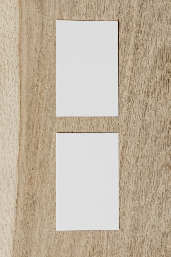 La imagen muestra dos tarjetas rectangulares en blanco colocadas sobre una superficie de madera con un patrón distintivo de vetas de madera. Las tarjetas están alineadas verticalmente con un espacio entre ellas, y la de arriba está ligeramente desplazada hacia la izquierda en comparación con la de abajo.