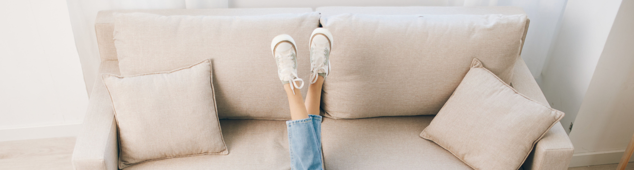 Pies vistiendo unas zapatillas blancas estilo Converse están cruzados sobre un sofá de color muy claro.