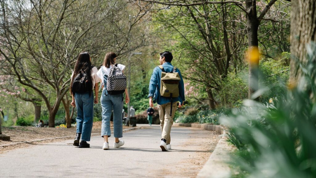 personas caminan por un sendero en un parque con abundante vegetación. Dos de ellas caminan juntas a la izquierda, una lleva una mochila y la otra tiene el cabello largo sobre el hombro. A la derecha, una persona con chaqueta azul y mochila amarilla camina un poco más adelante. Al fondo, se ven árboles con flores rosadas, sugiriendo que es primavera. El ambiente parece tranquilo y agradable, destacando el disfrute de la naturaleza y la compañía.