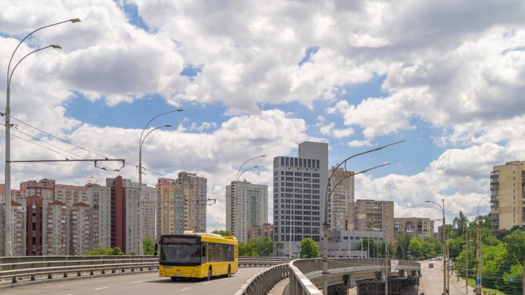 Una ciudad con un puente o paso elevado en primer plano. Un autobús amarillo circula por la carretera, y al fondo se ven varios edificios residenciales y comerciales. El cielo está parcialmente nublado, sugiriendo un día con sol y nubes intermitentes.