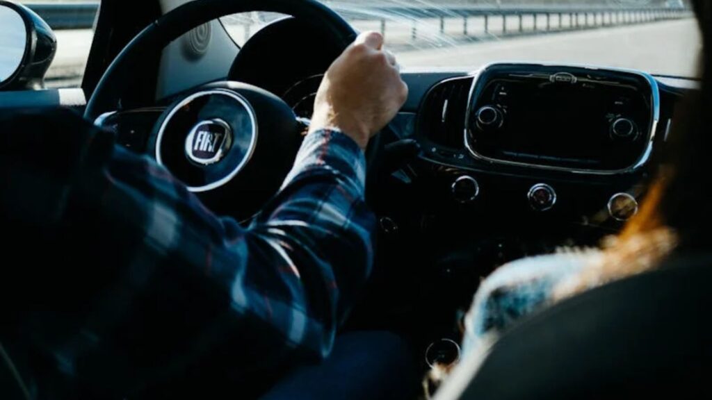 El interior de un vehículo visto desde la perspectiva de un pasajero en el asiento trasero. Se ve el volante con un emblema en el centro. La mano del conductor está en el volante y se ve parte de otro pasajero en primer plano a la derecha, lo que sugiere que hay al menos dos personas en el coche. El salpicadero contiene varios controles y un sistema de infoentretenimiento central.