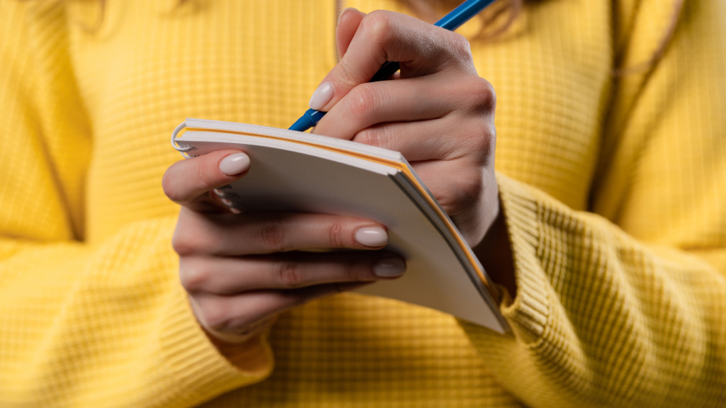 Primer plano de las manos de una persona sosteniendo un cuaderno pequeño abierto y escribiendo con un bolígrafo azul. La persona lleva un suéter amarillo de punto, y solo se ven las manos y parte del suéter en la imagen. El enfoque en la acción de escribir en el cuaderno hace que esta imagen sea relevante para temas relacionados con la toma de notas, el estudio, el diario o la planificación.