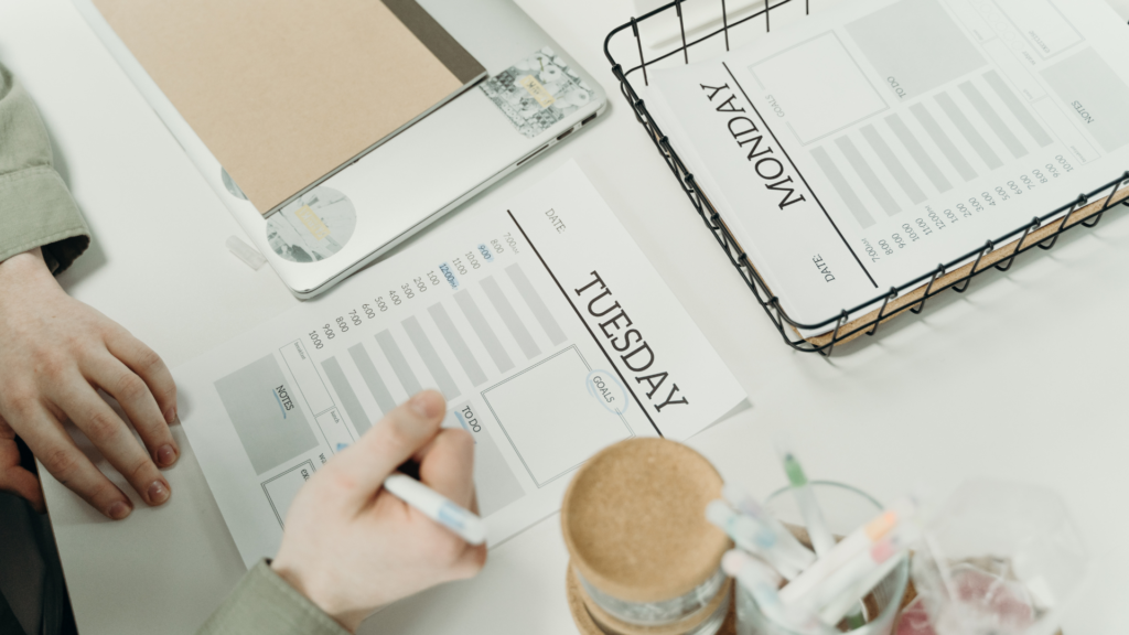 Manos de una persona escribiendo en una agenda abierta en una página etiquetada como “Tuesday”. La agenda está sobre un escritorio blanco, rodeada de varios objetos, incluyendo otro cuaderno, un teléfono móvil, un frasco de vidrio con utensilios de escritura y un pequeño contenedor con corcho. Esta imagen puede ser relevante para ilustrar la organización, actividades de planificación o conceptos de gestión del tiempo.