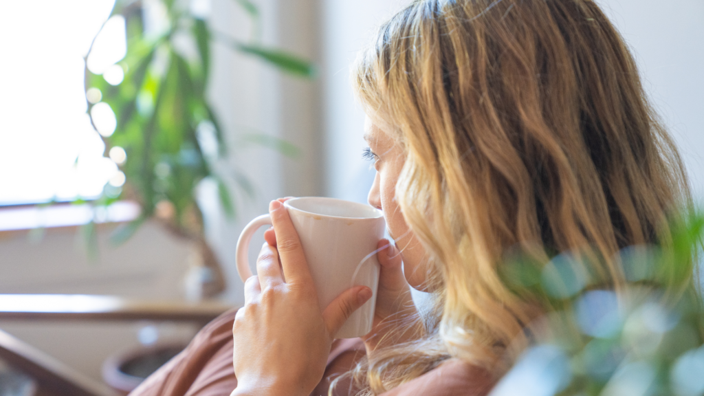 Persona con el rostro oculto sosteniendo una taza blanca, sentada en un entorno interior con un fondo desenfocado que presenta follaje verde y una ventana, lo que sugiere un ambiente relajado o doméstico para Recuperar el Orden Tras las Vacaciones.