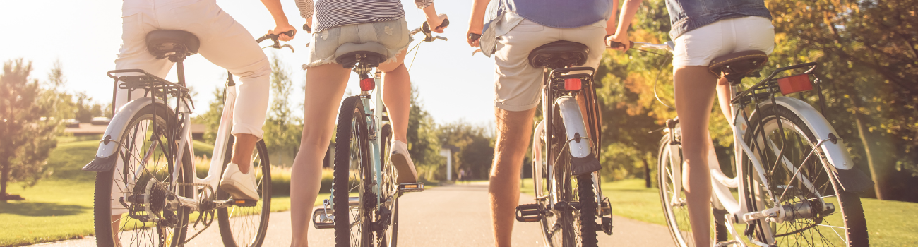 Cuatro personas montan en bicicleta por un sendero en un parque o área recreativa. La imagen está tomada desde atrás, enfocándose en la parte inferior de sus cuerpos y las bicicletas en movimiento. El entorno muestra un cielo despejado y vegetación exuberante a ambos lados del camino, sugiriendo un día agradable para actividades al aire libre.