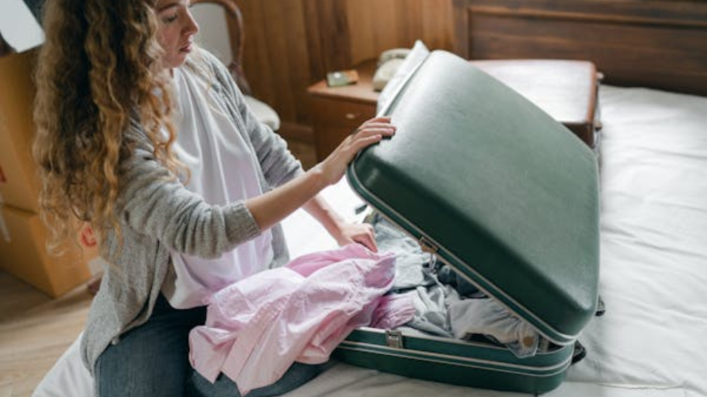 Persona con cabello largo y rizado desempacando una maleta verde con varias prendas, incluyendo ropa de color rosa. La maleta está sobre una cama y hay cajas de cartón en el fondo, lo que sugiere que la persona podría estar en proceso de mudanza o preparándose para un viaje.