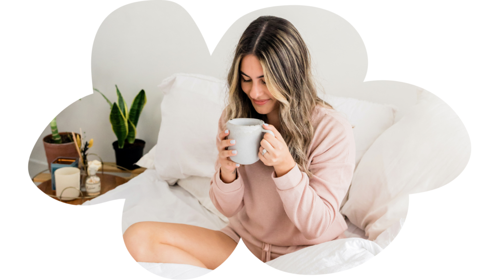 Persona sentada en una cama con ropa de cama blanca, sosteniendo una taza blanca. La persona lleva una camiseta rosa y está en una postura relajada con una pierna doblada bajo la otra. A la izquierda hay una mesita de noche de madera con varios objetos, incluyendo una planta pequeña, libros y posiblemente productos de cuidado personal. El fondo es minimalista y principalmente blanco, lo que resalta al sujeto.