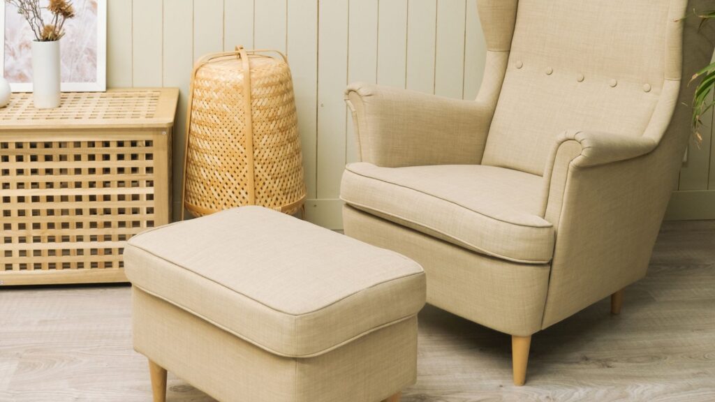 Un sillón tapizado en beige con un reposapiés a juego, colocado sobre un suelo de madera clara frente a una pared amarilla pálida. A la izquierda del sillón hay un cesto de ratán que sirve también de mesa auxiliar, o sea un sistemas de almacenamiento multifuncional, y un pequeño aparador con un jarrón pequeño y plantas secas, creando un ambiente acogedor y minimalista.