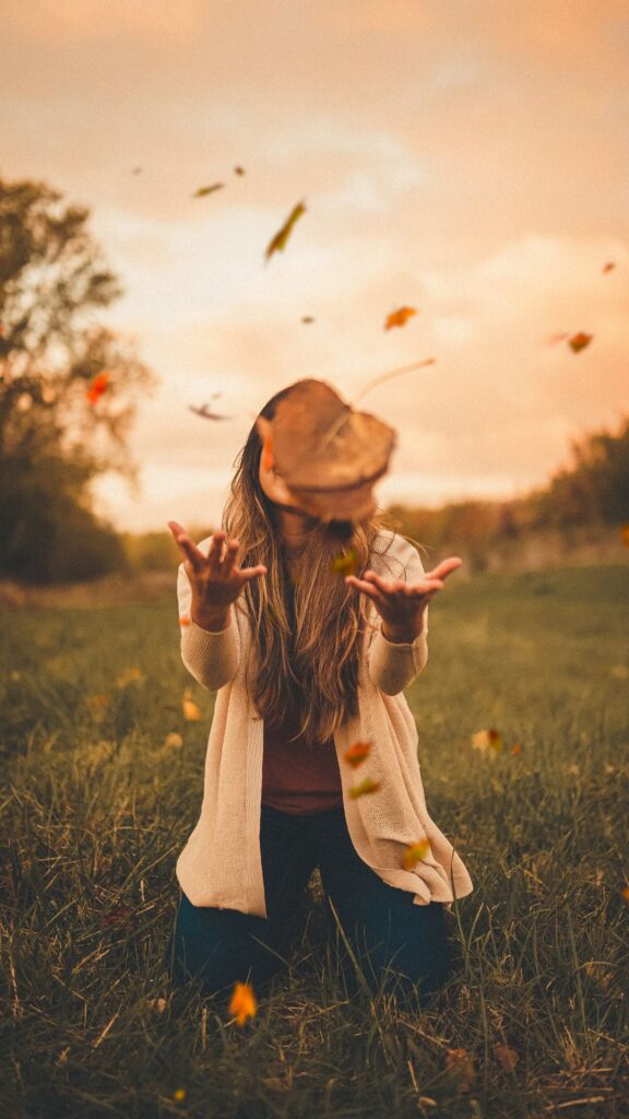 La imagen muestra a una persona en un campo con hojas de otoño cayendo a su alrededor. La persona tira las hojas hacia arriba y las coge en el aire. Su rostro está oculto por sus manos extendidas para coger las hojas, como si estuviera jugando con ellas. La escena transmite una sensación de movimiento y diversión otoñal.