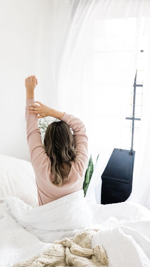 La imagen muestra a una persona sentada en una cama con ropa de cama blanca y estirándose con un brazo extendido hacia arriba. La persona lleva una camiseta de manga larga rosa y tiene el cabello oscuro. En el fondo, hay una ventana con cortinas translúcidas, a través de las cuales entra luz natural. Esta escena podría ser interesante o relevante ya que representa un momento de relajación o rutina matutina en un entorno de dormitorio sereno.