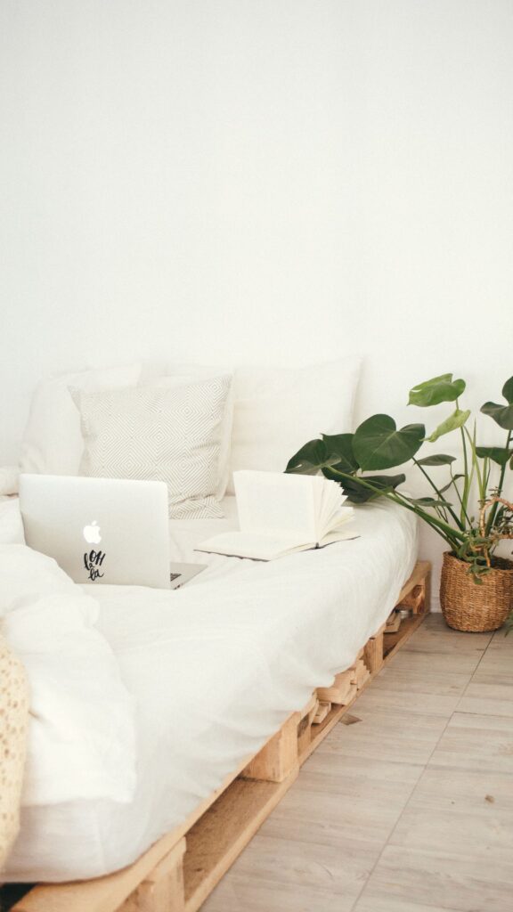 Un dormitorio con una cama hecha de madera de palets, ropa de cama blanca y un portátil abierto sobre la cama. Al lado de la cama hay una planta en maceta con hojas grandes y verdes, y un cesto de mimbre en el suelo. El diseño es minimalista con colores neutros.