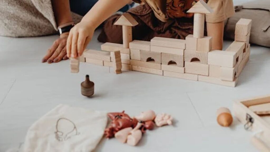 Una estructura de bloques de madera que forma un castillo con múltiples torres y arcos, rodeada de juguetes y bloques sueltos, con las manos de una persona ajustando una de las piezas.