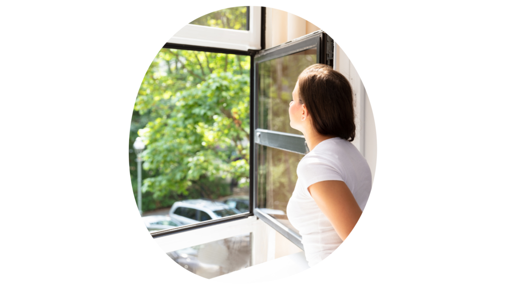 Mujer mirando hacia un árbol desde la ventana abierta y respirando el aire que entra.