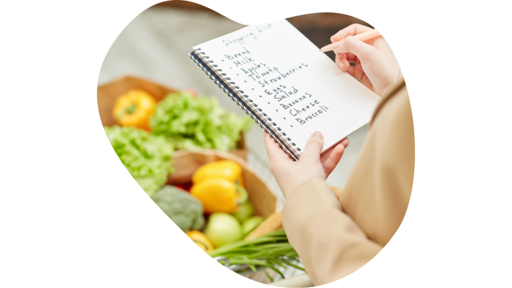 Mano femenina anotando en un cuaderno una lista de la compra y al lado hay una bolsa de papel con frutas y verduras.