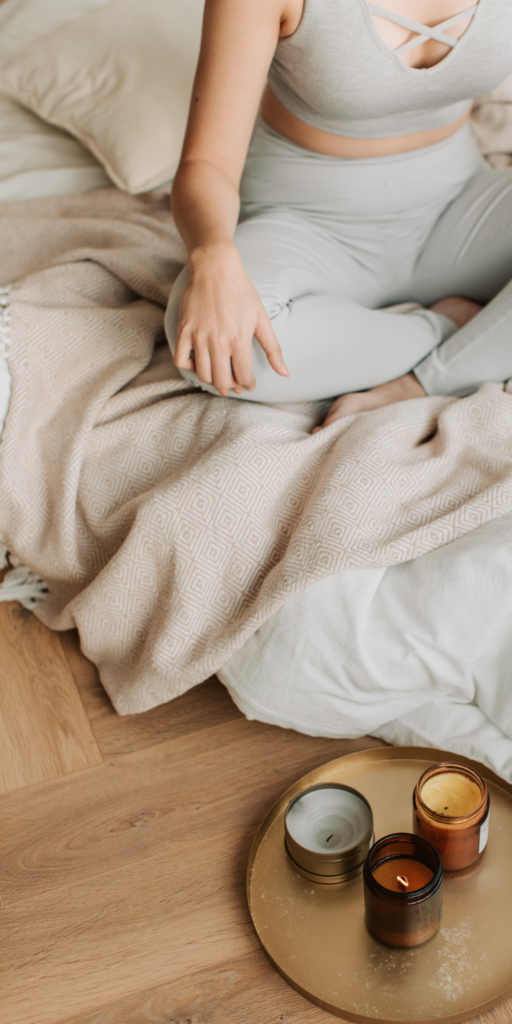 Una persona sentada en una cama con ropa de cama beige, usando leggings grises claros y un top corto blanco. Frente a la persona hay una bandeja redonda con tres velas encendidas de diferentes tamaños y colores, creando una atmósfera propicia para la aromaterapia y la meditación. La escena evoca una sensación de calma y relajación, esenciales en las prácticas de aromaterapia y meditación.