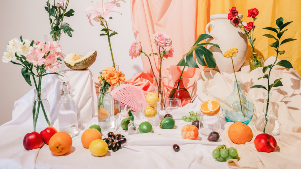 Una vibrante composición de naturaleza muerta sobre un mantel blanco. La escena incluye una variedad de flores coloridas en diferentes tipos de jarrones y botellas, algunas de vidrio transparente y otras de vidrio coloreado. También hay diversas frutas como uvas, naranjas, limones y una papaya cortada por la mitad, junto con follaje verde intercalado. Un abanico rosa está parcialmente abierto cerca del centro. El fondo consiste en telas drapeadas en tonos de naranja y rosa, que complementan la paleta de colores cálidos de la composición.