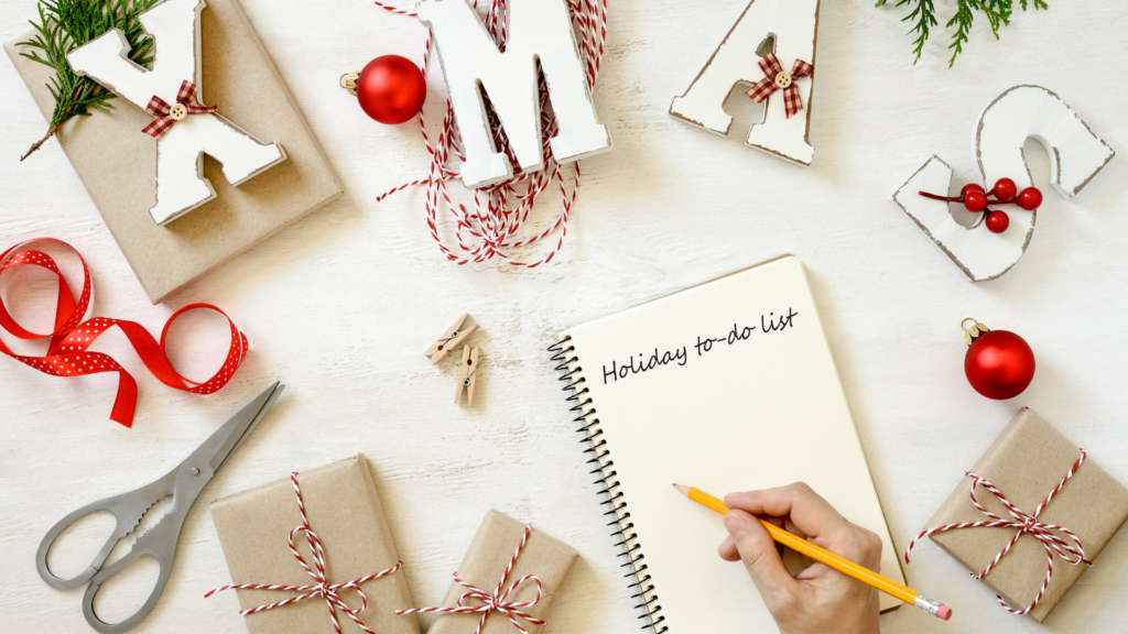 La imagen muestra un espacio de trabajo festivo con varias decoraciones navideñas y materiales para envolver regalos. Hay regalos envueltos con cuerda roja y blanca, cinta roja, tijeras y letras de madera que forman la palabra "XMAS". Una mano sostiene un lápiz y escribe en un bloc de notas con el encabezado "Holiday to-do list". La escena es festiva y organizada, lo que la hace relevante para la planificación de una Navidad consciente y la preparación de regalos.