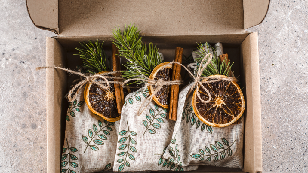 La imagen muestra una caja de cartón que contiene tres pequeñas bolsas de tela. Cada bolsa está decorada con un patrón de hojas verdes y atada con un trozo de cuerda. Adjuntos a la cuerda en cada bolsa hay una rodaja de naranja seca, una rama de canela y una ramita de pino. Esta imagen es interesante y relevante ya que representa un regalo o decoración rústica y natural, probablemente destinada a una ocasión festiva o especial. La combinación de rodajas de naranja secas, ramas de canela y ramitas de pino sugiere una presentación festiva, aromática y visualmente atractiva.