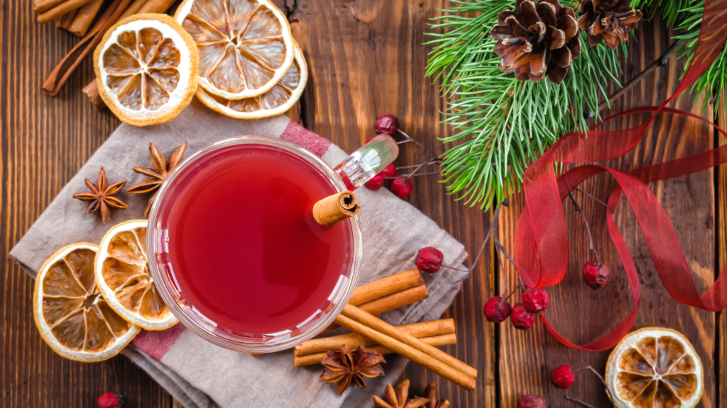 La imagen muestra una taza de vidrio llena de una bebida roja, probablemente vino caliente o sidra especiada, con una rama de canela dentro. Alrededor de la taza hay varios elementos temáticos de la Navidad, como rodajas de naranja secas, anís estrellado, ramas de canela, piñas, ramas de pino, bayas rojas y una cinta roja. El escenario está sobre una superficie de madera, lo que añade un ambiente rústico y acogedor, evocando una sensación cálida y festiva, perfecta para una Navidad consciente.